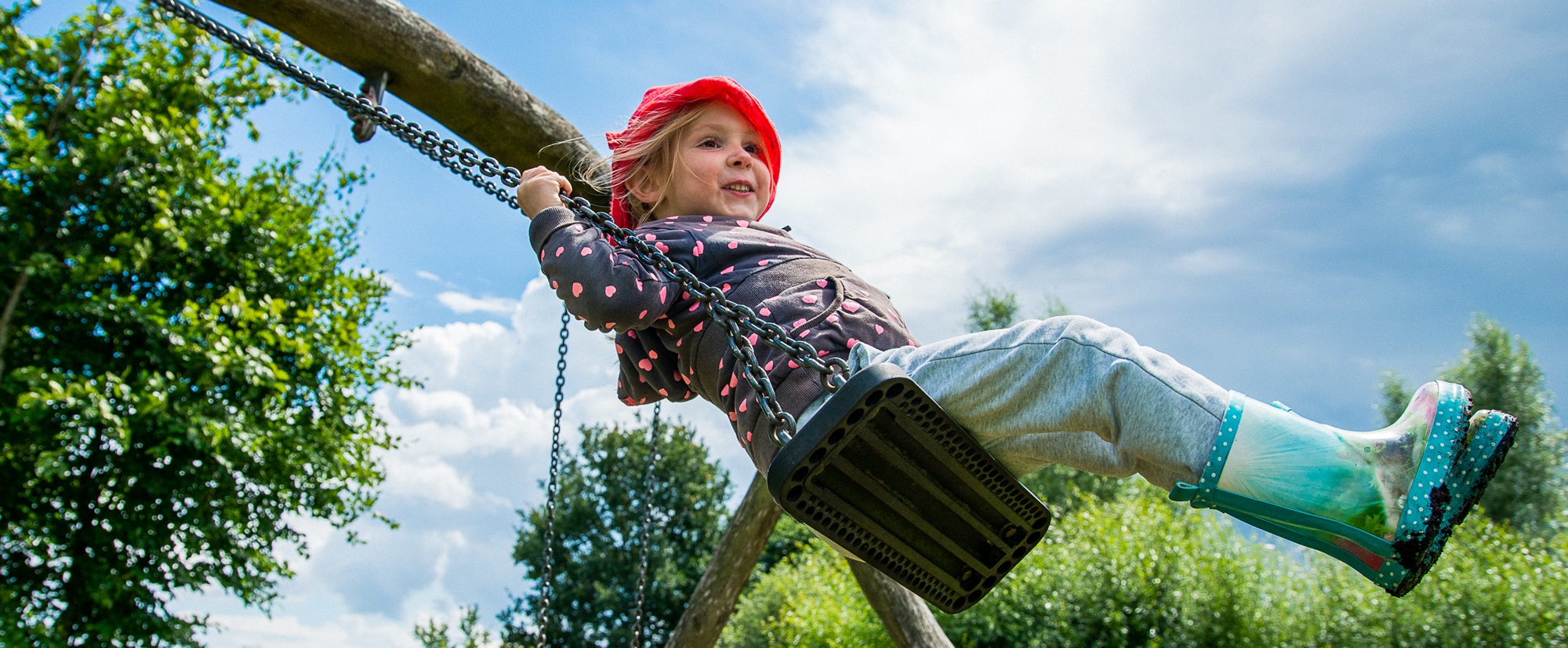 Een meisje met een rode kaboutermuts schommelt in een buitenspeeltuin tijdens een middag vol kinderactiviteiten in het Hart van Limburg.