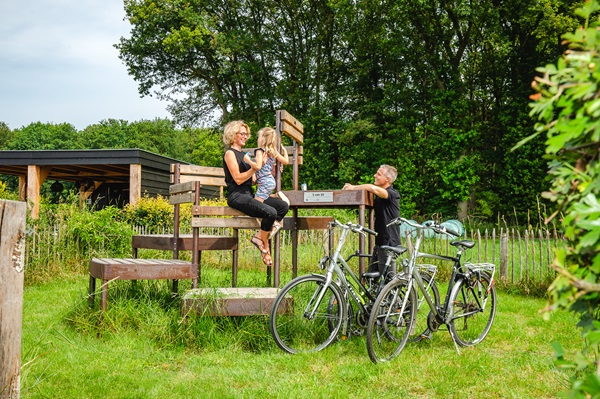 Een gezin zit op een houten kunstwerk in een groene omgeving, met fietsen op de voorgrond.