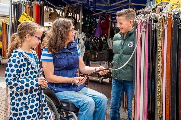 Drie mensen bekijken en bespreken een riem bij een marktkraam op de weekmarkt in Weert. Een vrouw in een rolstoel houdt de riem vast, terwijl een jongen en een meisje toekijken en lachen. Op de achtergrond zijn marktkramen met tassen en accessoires te zien.