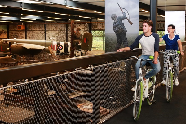 Fietsers kijken vanaf de fietsersbrug in het Oorlogsmuseum Overloon naar de bezienswaardigheden