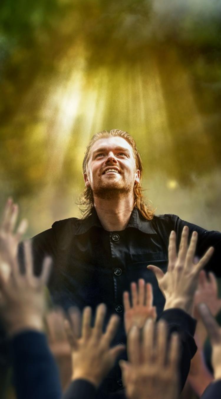 Man met uitgestrekte armen in een bosachtig decor, terwijl zonlicht op hem valt en handen uit het publiek naar hem reiken tijdens de Passiespelen in Tegelen.